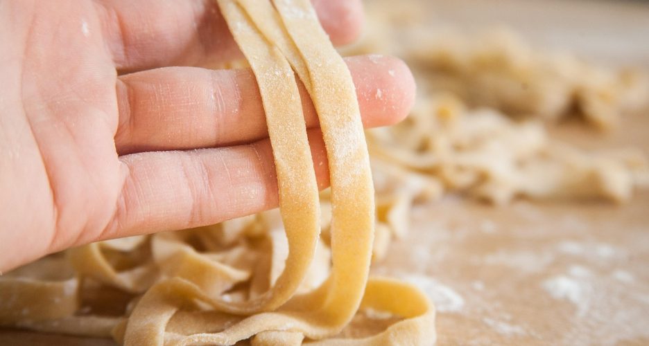 Dłoń formująca świeży makaron tagliatelle z ciasta rozłożonego na blacie z mąką.
